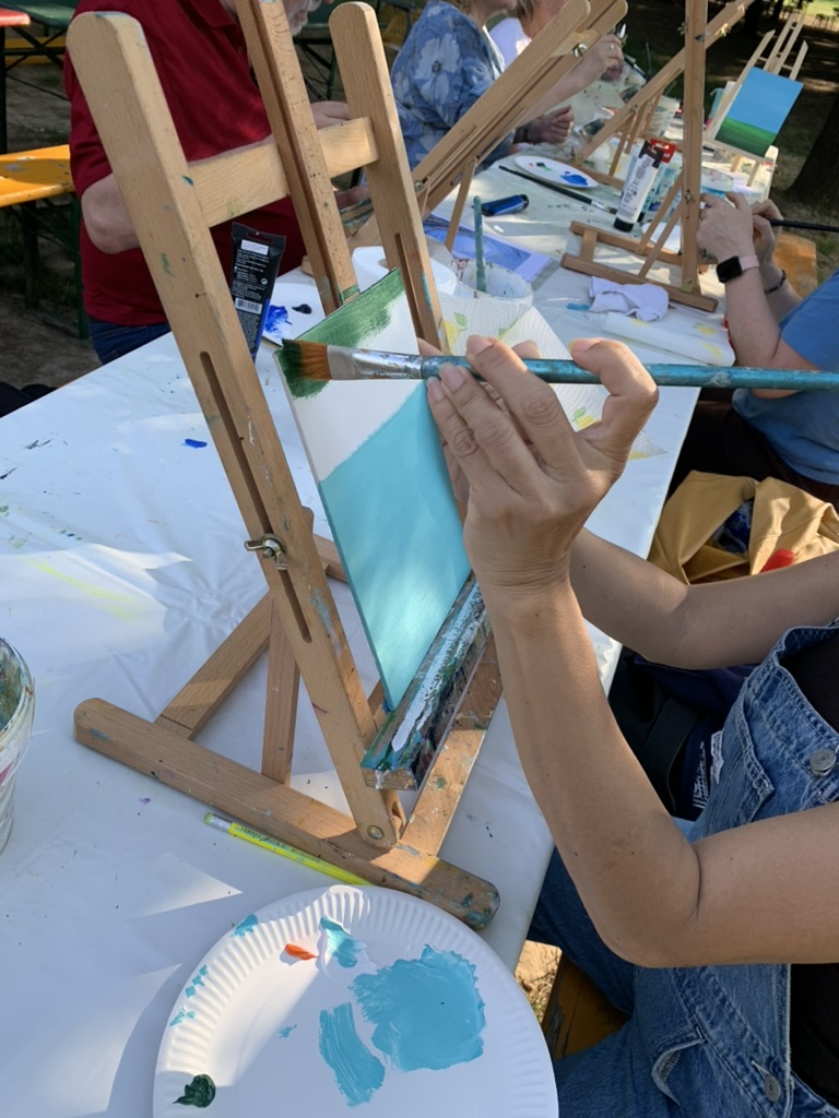 Mani che dipingono di turchese una piccola tela sul cavalletto durante un workshop di pittura all'aperto